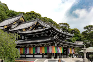 A traditional Japanese temple with ornate black-and-white tiled roofs and colorful fabric banners hanging along the entrance, surrounded by lush green trees under a partly cloudy sky.
