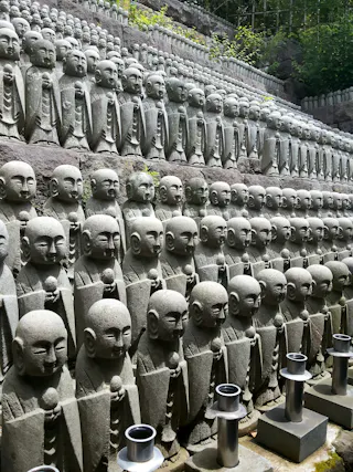 Rows of stone statues of Buddhist monks are neatly arranged on tiered steps outdoors, surrounded by greenery. Each statue holds an object and has a serene expression. Metal candle holders are placed in front of some statues.