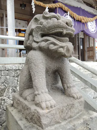 A stone guardian lion-dog statue (shisa or komainu) sits at the entrance of a traditional Japanese building with purple and white banners and rope decorations in the background.