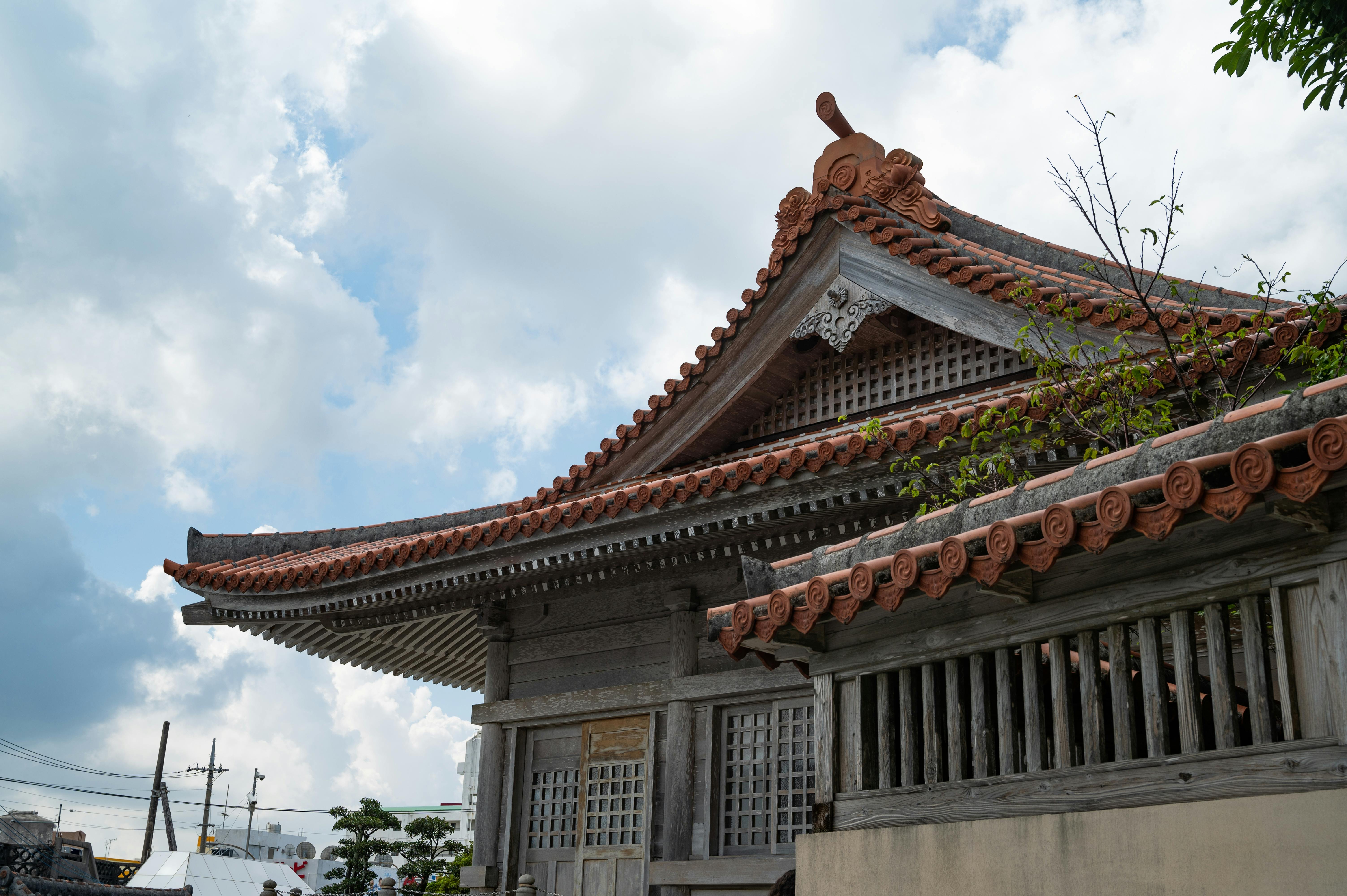 A traditional Japanese building with a decorative, tiled roof and wooden architecture under a partly cloudy sky, with trees and power lines visible in the background.