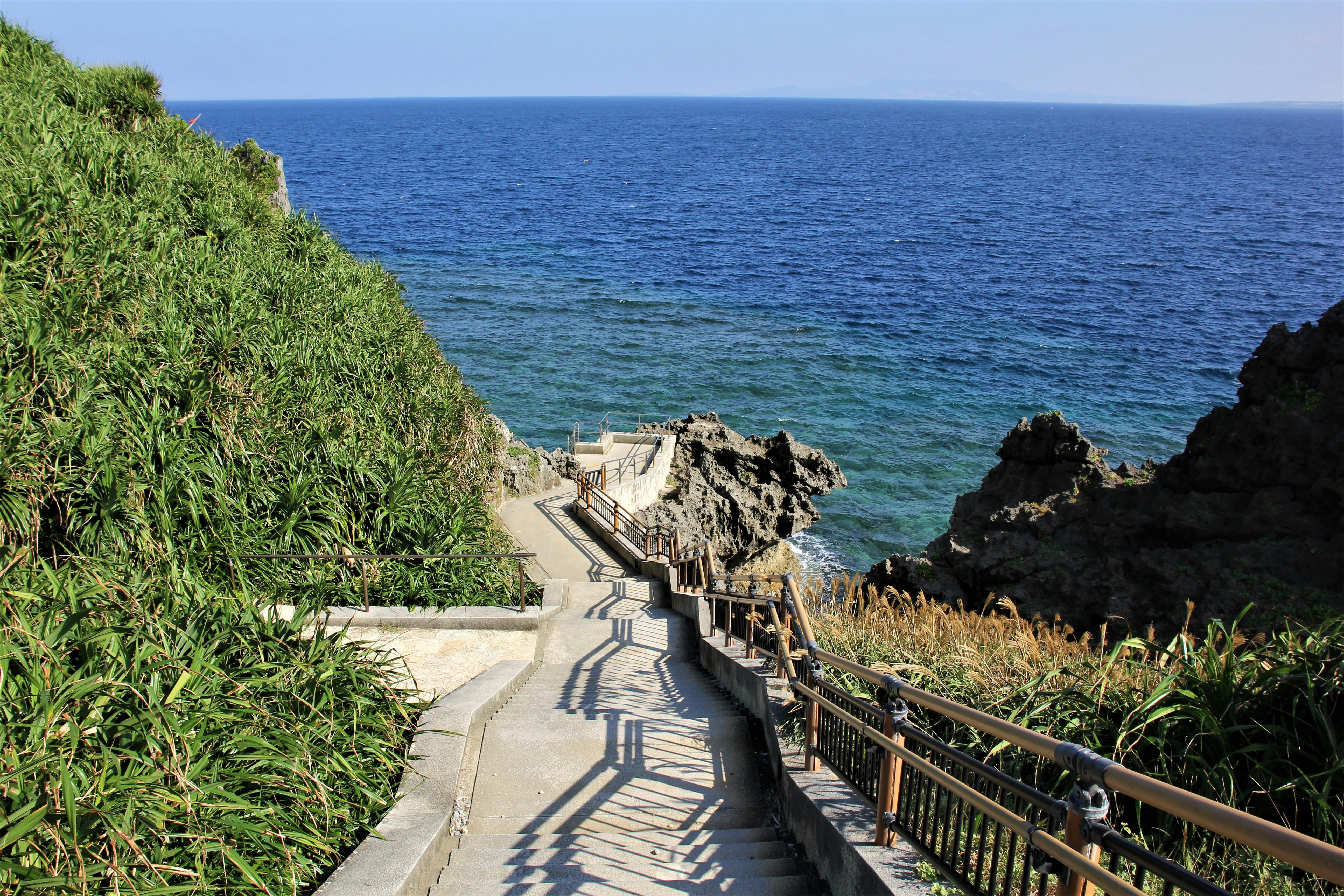 A concrete pathway with railings descends between green bushes and rocky cliffs toward the blue ocean under a clear sky. The stairs cast shadows along the walkway leading to the rocky shoreline.