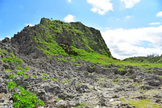 Rocky hill covered with patches of green vegetation under a bright blue sky with scattered clouds. The rough, jagged terrain dominates the foreground, with sunlight highlighting the natural landscape.