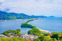 Aerial view of Amanohashidate in Japan, showing a sandbar covered with green pine trees stretching across blue water, with mountains and a coastal town in the background under a partly cloudy sky.