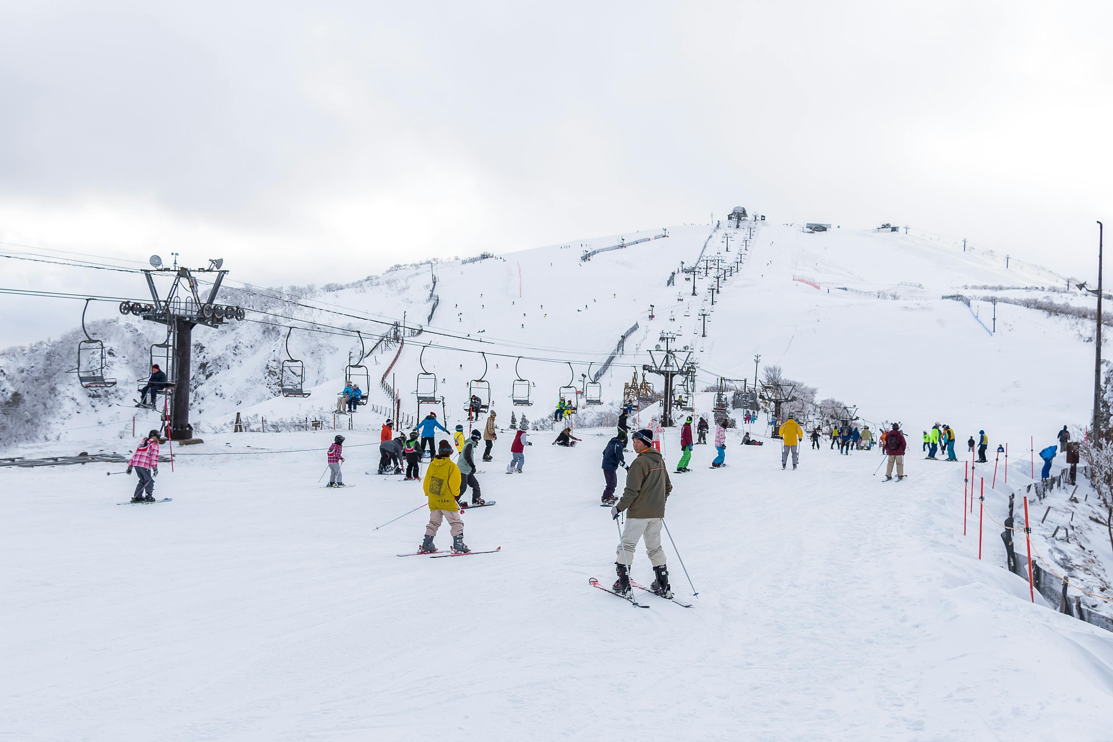 Skiers and snowboarders enjoy a snowy ski resort, with chairlifts in operation and people gathered on the slope. Snow-covered hills and cloudy skies are visible in the background.