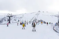 Skiers and snowboarders enjoy a snowy ski resort, with chairlifts in operation and people gathered on the slope. Snow-covered hills and cloudy skies are visible in the background.