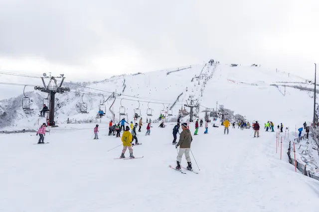 Skiers and snowboarders enjoy a snowy ski resort, with chairlifts in operation and people gathered on the slope. Snow-covered hills and cloudy skies are visible in the background.