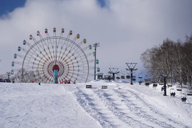 A snowy ski slope with people skiing and riding chairlifts. In the background, there is a large Ferris wheel with colorful gondolas. Leafless trees border the right side of the scene under a partly cloudy sky.