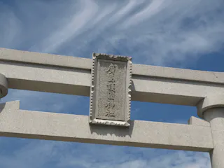 A close-up of the top section of a traditional stone torii gate with Japanese kanji inscribed on a plaque, set against a blue sky with wispy white clouds.