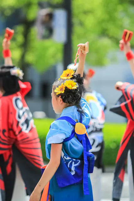 A group of people in colorful traditional Japanese clothing perform an outdoor dance. The central person has flowers in their hair and raises a small wooden instrument with one hand. Lush green trees are visible in the background.