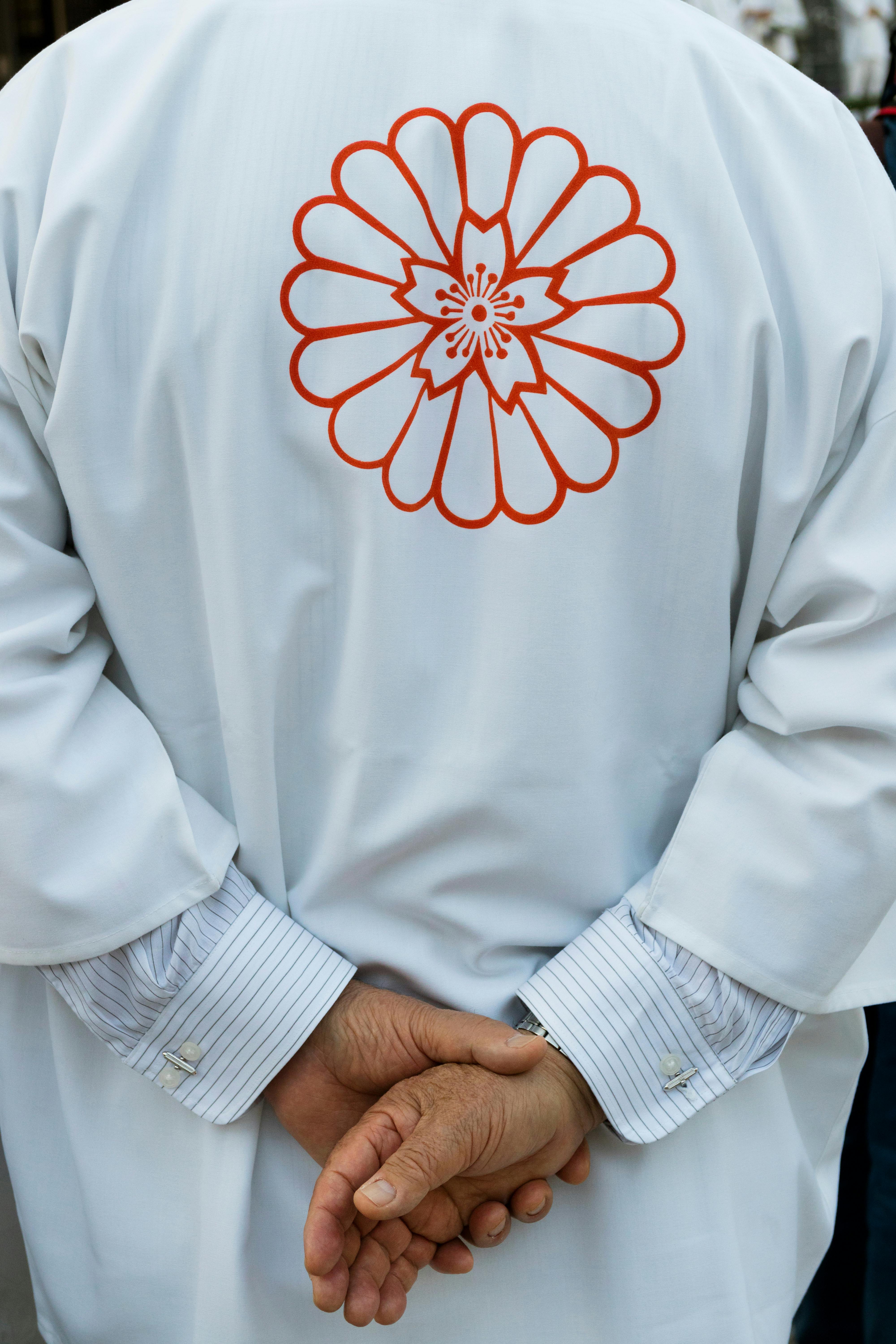 A person stands with hands clasped behind their back, wearing a white garment featuring a large red chrysanthemum flower design on the back.