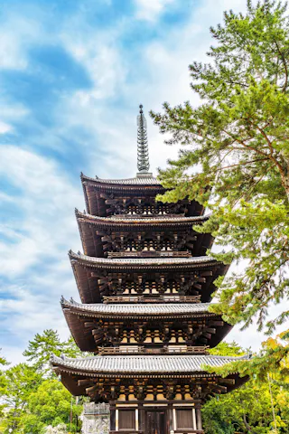 A traditional five-story wooden pagoda stands surrounded by green trees under a partly cloudy blue sky.