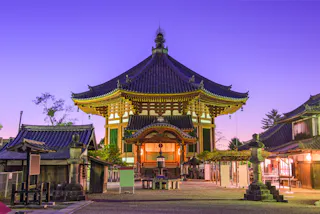 A traditional Japanese temple with ornate architecture is illuminated at dusk, surrounded by lanterns and smaller buildings, against a purple twilight sky.
