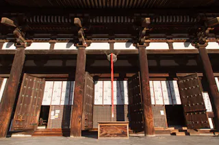 A traditional Japanese temple entrance with large wooden doors, tall wooden pillars, and a red and white rope hanging above a wooden offertory box in the sunlight.
