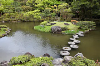 A tranquil Japanese garden with a pond, circular stone stepping stones leading to a grassy island with rocks and lush green trees surrounding the water.