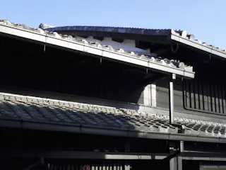 Traditional Japanese tiled rooftops with wooden beams and eaves are shown in strong sunlight, casting shadows on the dark exterior walls of the building against a clear blue sky.