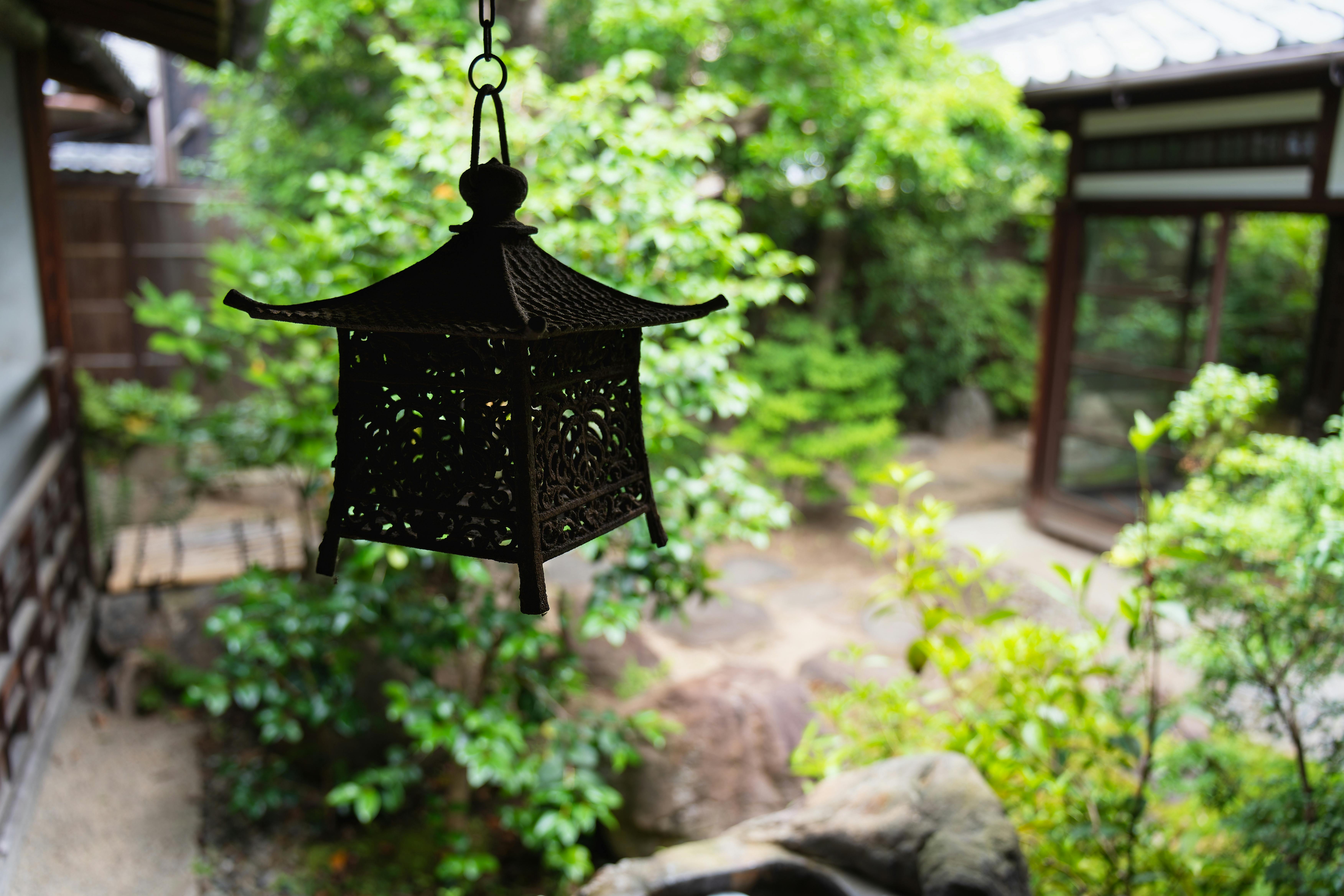 A black, ornate lantern hangs in the foreground of a tranquil Japanese garden, with lush green plants, rocks, and wooden structures visible in the soft focus background.