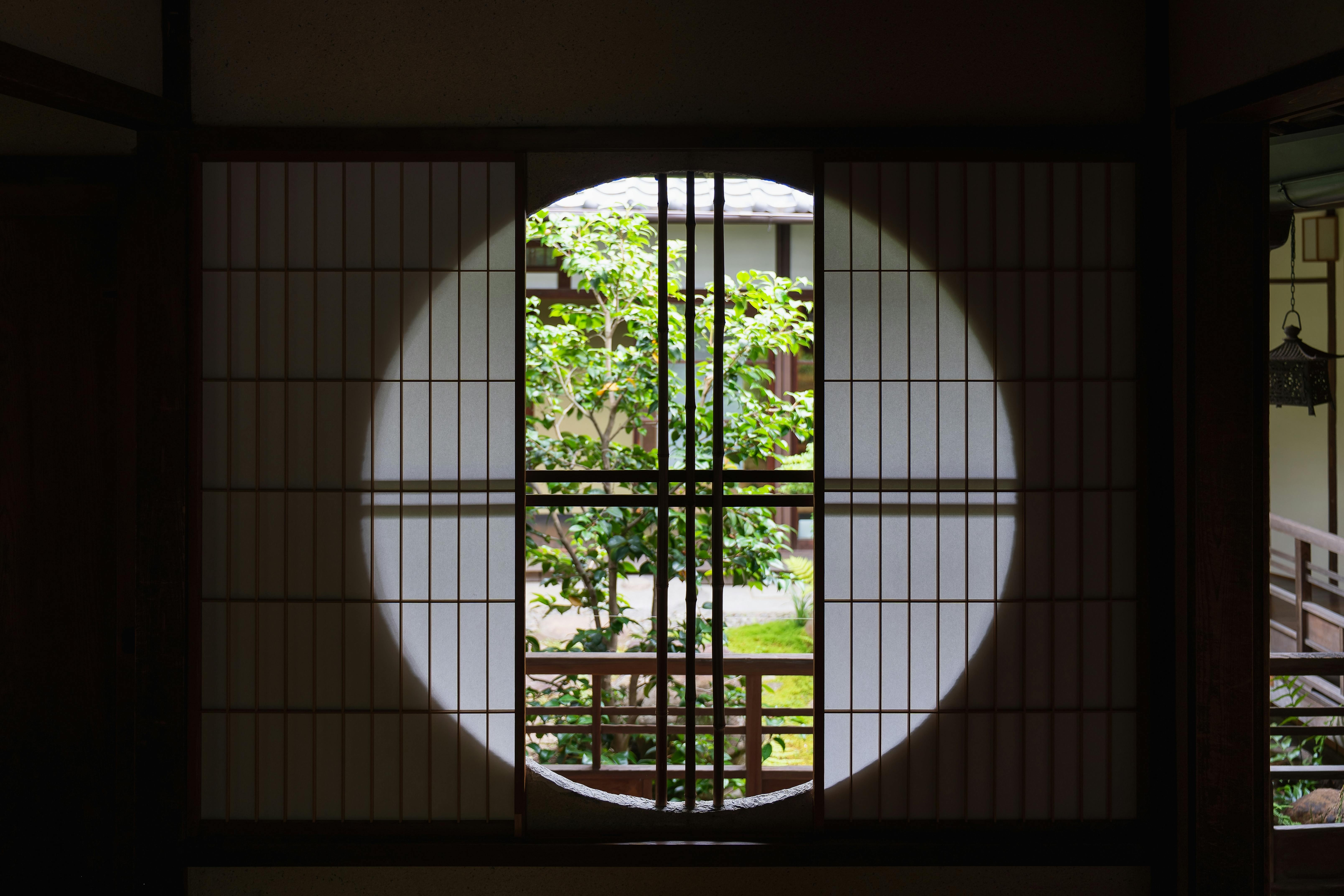 A round window framed by wooden latticework looks out onto a green garden with a leafy tree, set against traditional Japanese architecture in the background. Sunlight highlights the circular shape on the paper screen.