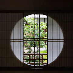 Naramachi A round window framed by wooden latticework looks out onto a green garden with a leafy tree, set against traditional Japanese architecture in the background. Sunlight highlights the circular shape on the paper screen.