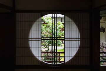 Naramachi A round window framed by wooden latticework looks out onto a green garden with a leafy tree, set against traditional Japanese architecture in the background. Sunlight highlights the circular shape on the paper screen.