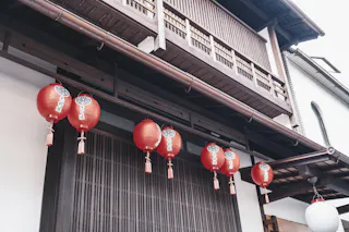 Red paper lanterns with Japanese characters hang in a row outside a traditional wooden building with sliding doors and wooden latticework, viewed from below.