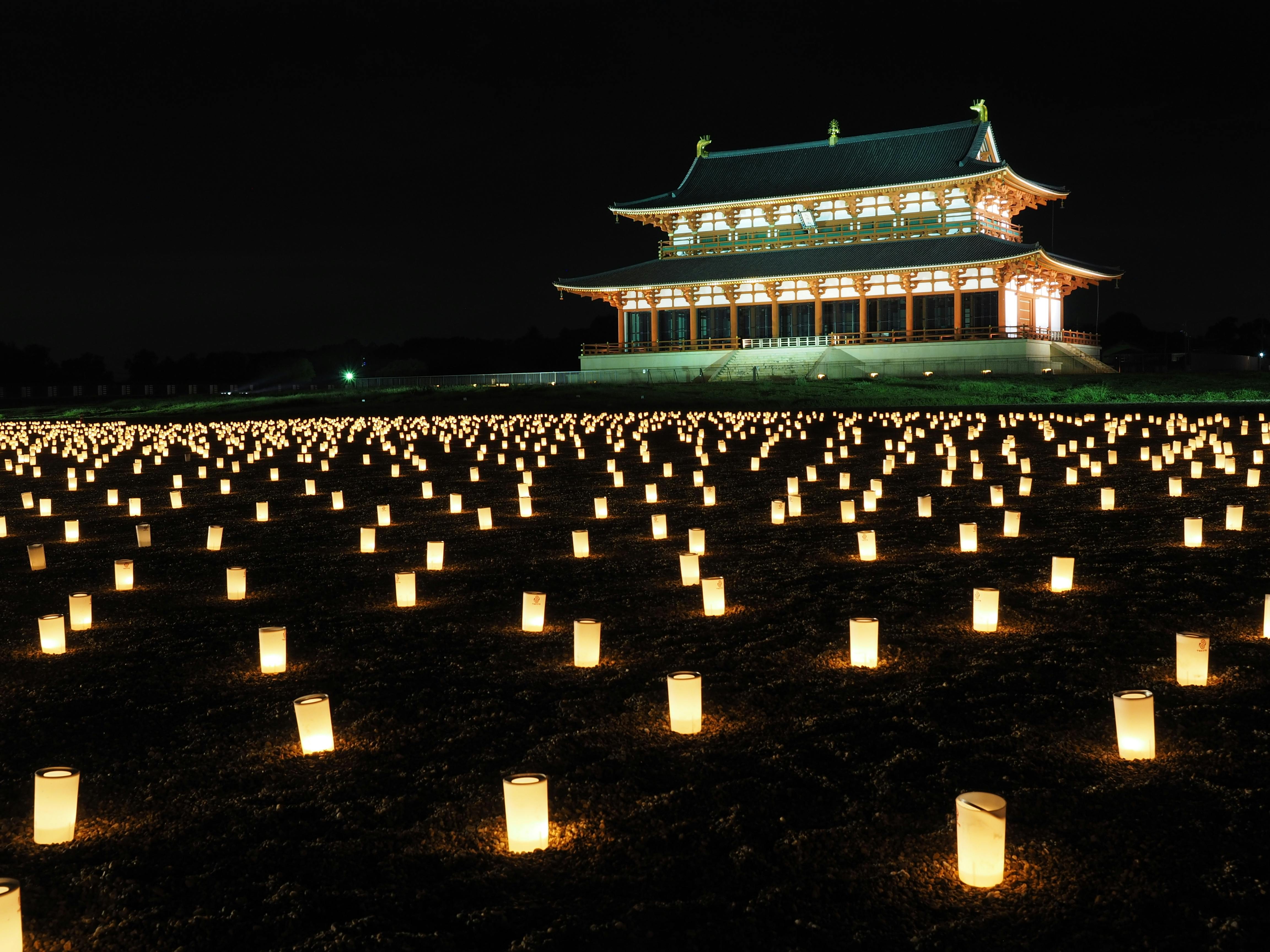 A traditional Japanese building illuminated at night, with hundreds of glowing lanterns arranged in rows on the ground in front of it, creating a serene and festive atmosphere.