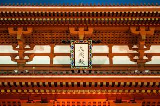 A close-up view of an ornate, wooden Japanese temple gate, featuring intricate patterns, gold accents, and a central sign with Japanese characters. The background shows a clear sky.