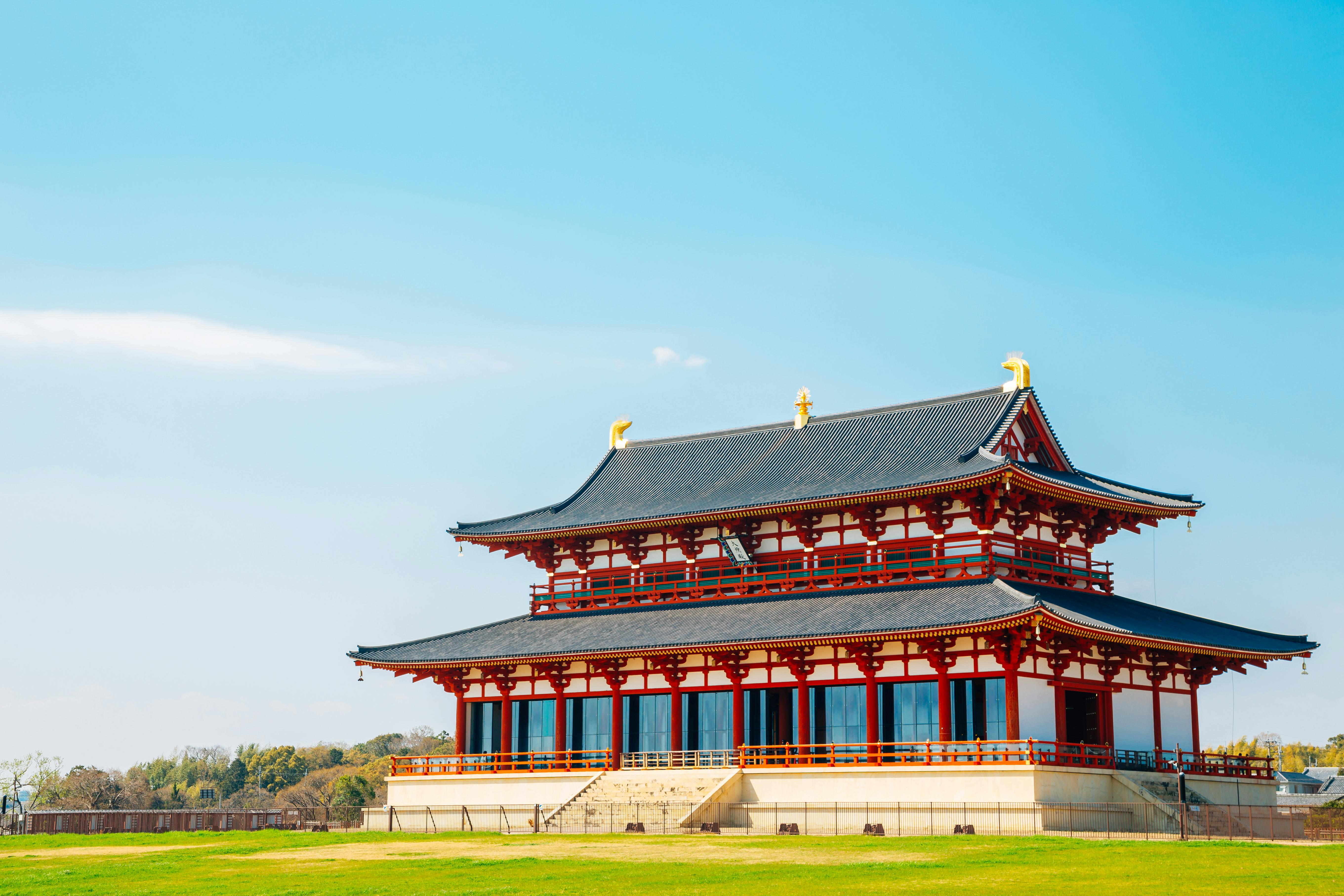 A traditional Japanese temple with a black tiled roof and red-and-white wooden beams stands on a grassy field under a clear blue sky.