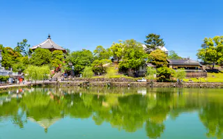 A peaceful pond reflects lush green trees and traditional Japanese buildings under a clear blue sky, creating a serene landscape.