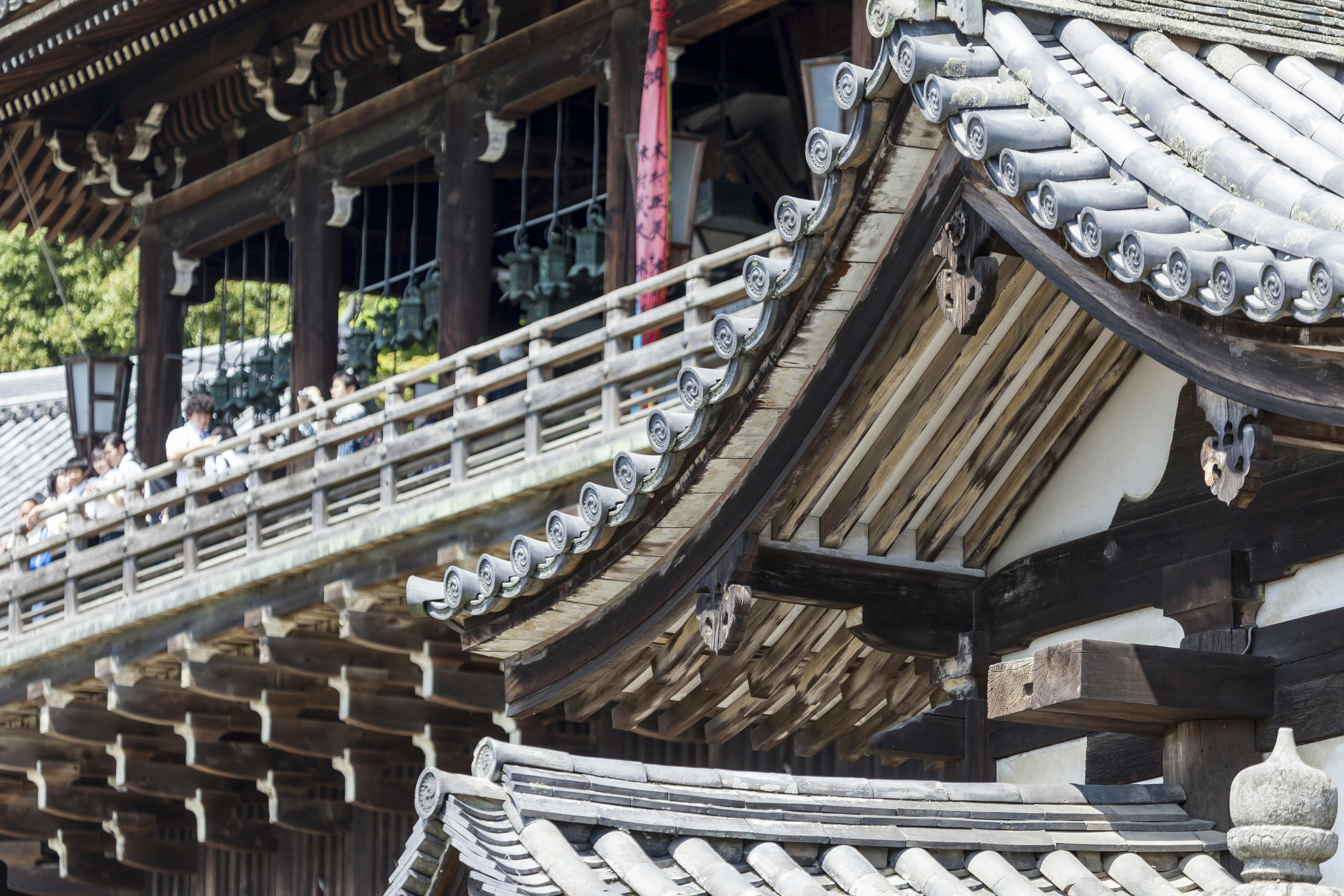 Traditional Japanese temple with curved tiled roofs and wooden beams, people standing on a balcony in the background, surrounded by detailed architectural elements and greenery.