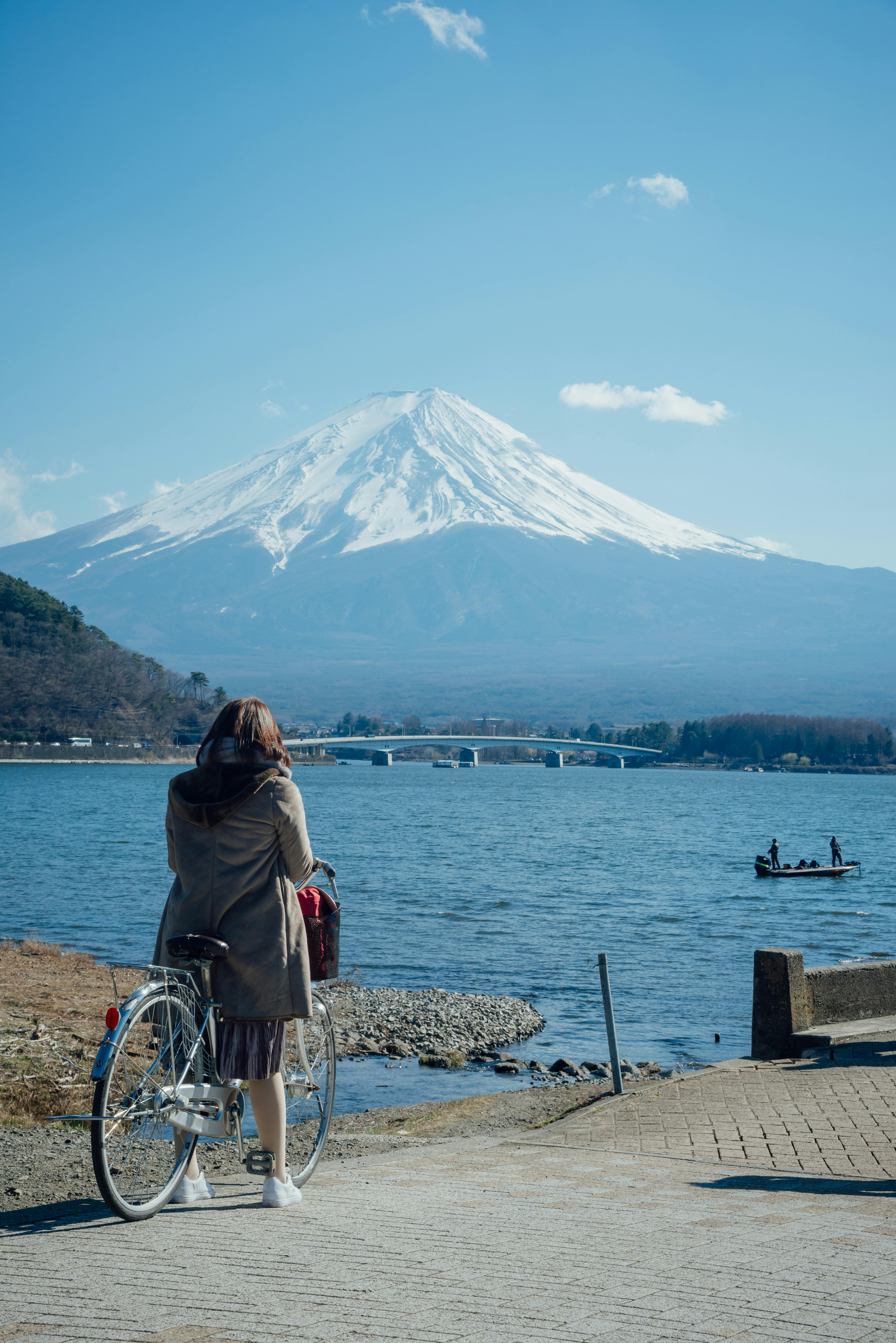 A person with a bicycle stands by a lakeshore, looking towards Mount Fuji in the distance under a clear blue sky. Two boats with people are visible on the lake.