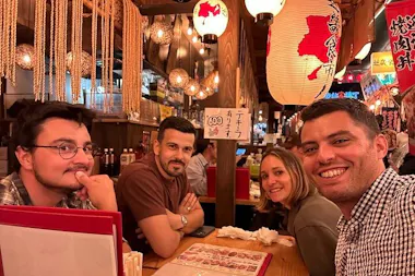 Four people sit at a cozy restaurant table, smiling at the camera. The setting is warmly lit with hanging lanterns, wooden decor, Japanese signs, and menus visible, creating a lively, inviting atmosphere.