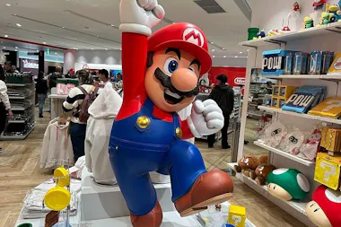 A large Mario statue with a raised fist stands in the center of a brightly lit Nintendo store, surrounded by merchandise, shelves with power-ups, and shoppers in the background.