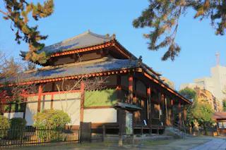 A traditional Japanese temple with a tiled roof and wooden beams stands surrounded by trees and plants on a sunny day, with modern buildings visible in the background.