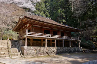 A traditional Japanese wooden temple with a sloped roof, raised on a stone foundation, stands beside stone steps, surrounded by trees and greenery.