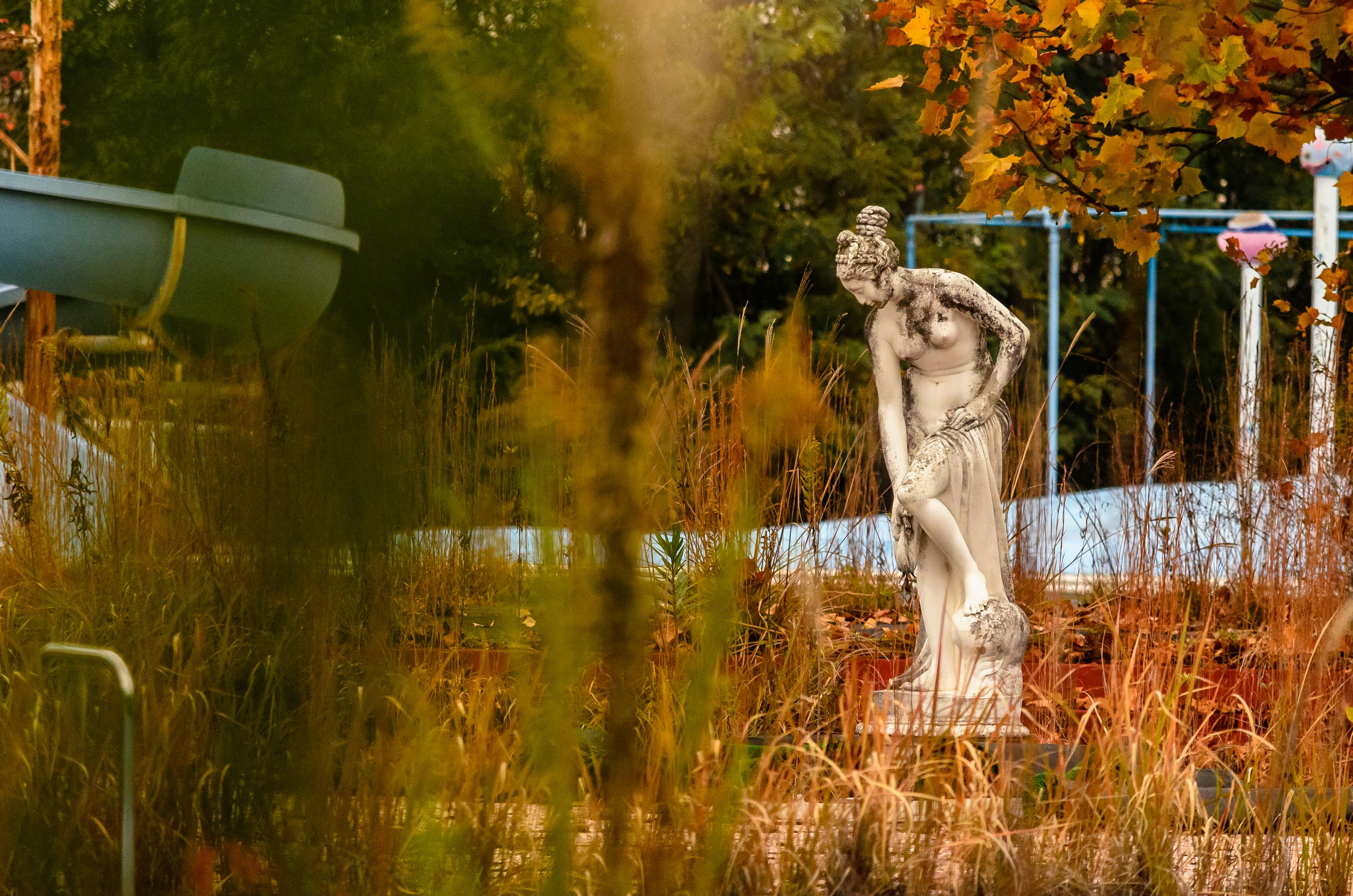 A white classical statue of a woman stands in an overgrown, abandoned outdoor area with tall grass, autumn leaves, and an empty waterslide in the background.