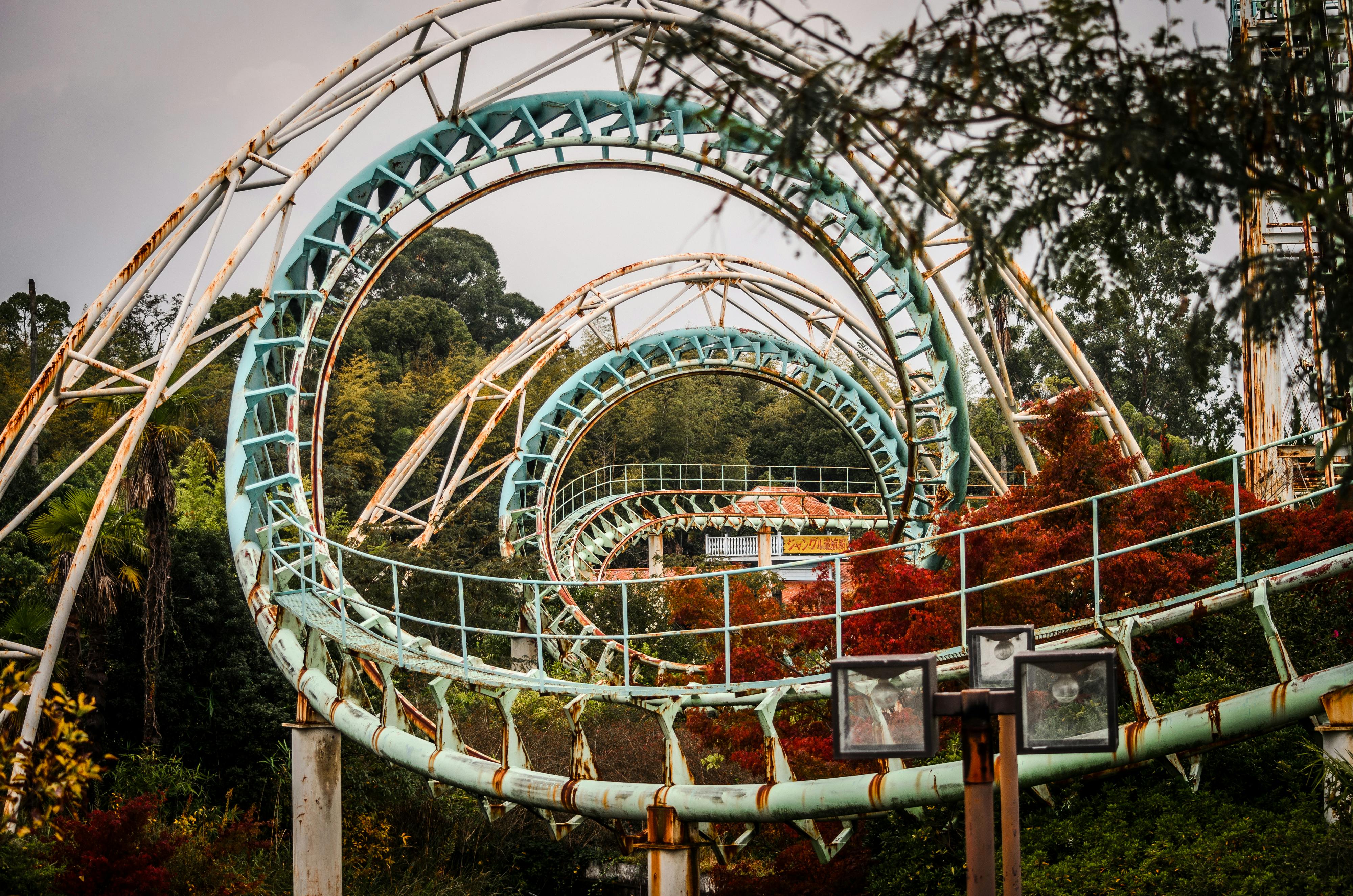 A rusty, abandoned roller coaster with teal loops winds through overgrown trees and foliage, showing signs of neglect and decay.