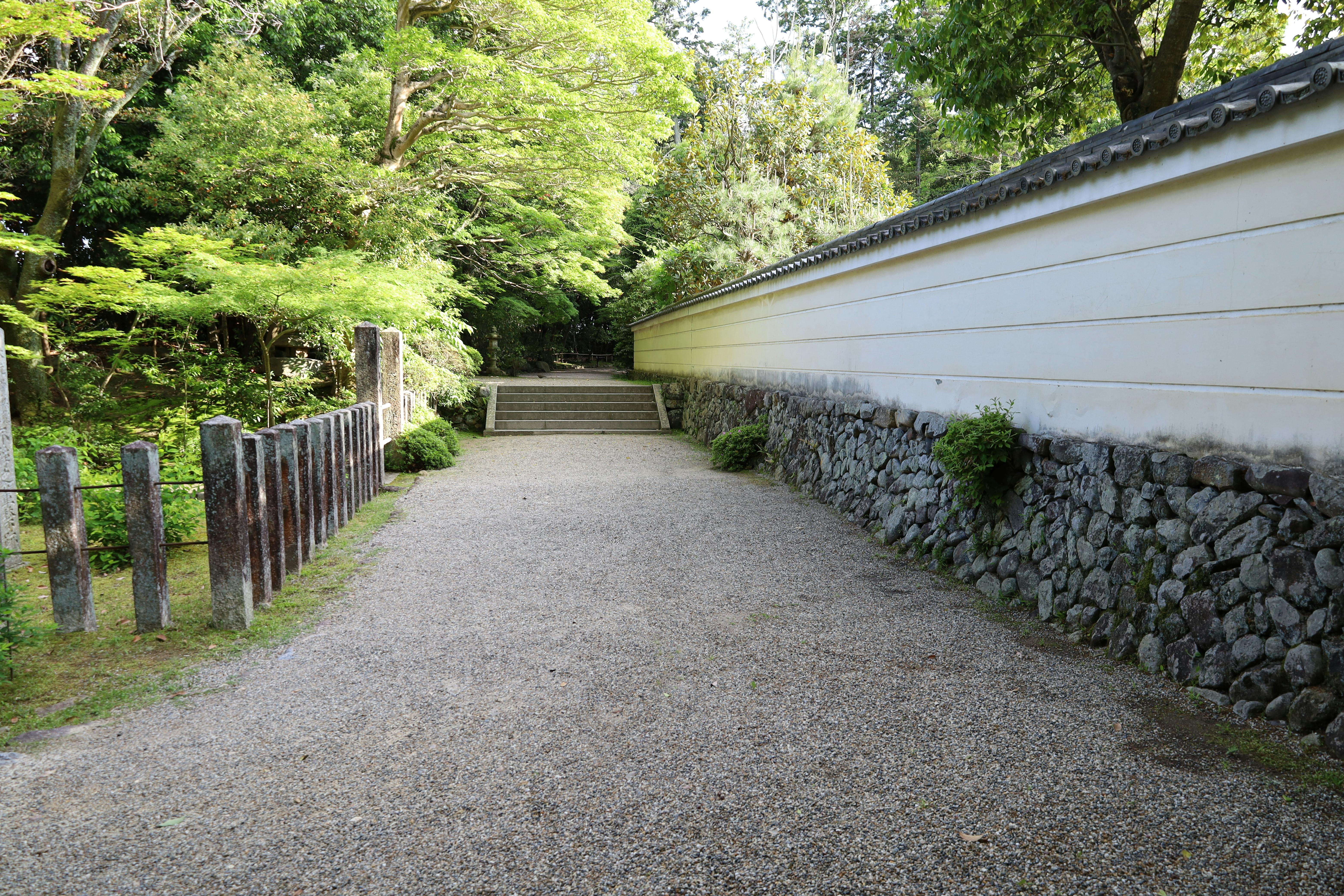 A gravel path bordered by a white stone wall and wooden posts curves through a lush, green garden with trees and shrubs, leading to stone steps in the background.