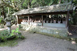 A wooden shrine structure with a tiled roof houses several small altars, surrounded by lush trees and a gravel path. A traditional stone lantern stands nearby on a mossy patch.
