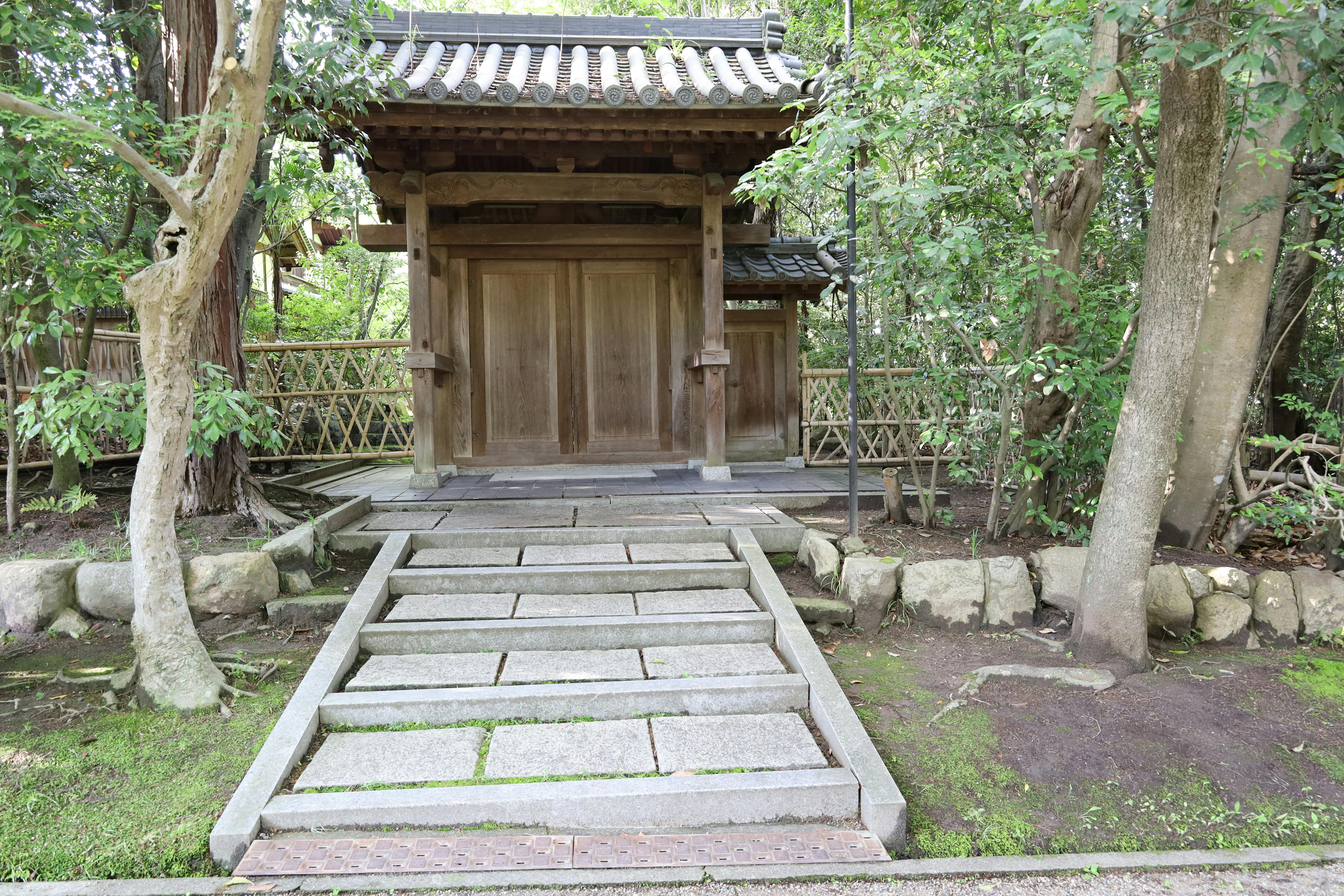 Traditional Japanese wooden gate with closed doors, flanked by lush green trees and stone pathway steps leading up to the entrance. A bamboo fence runs along the background.