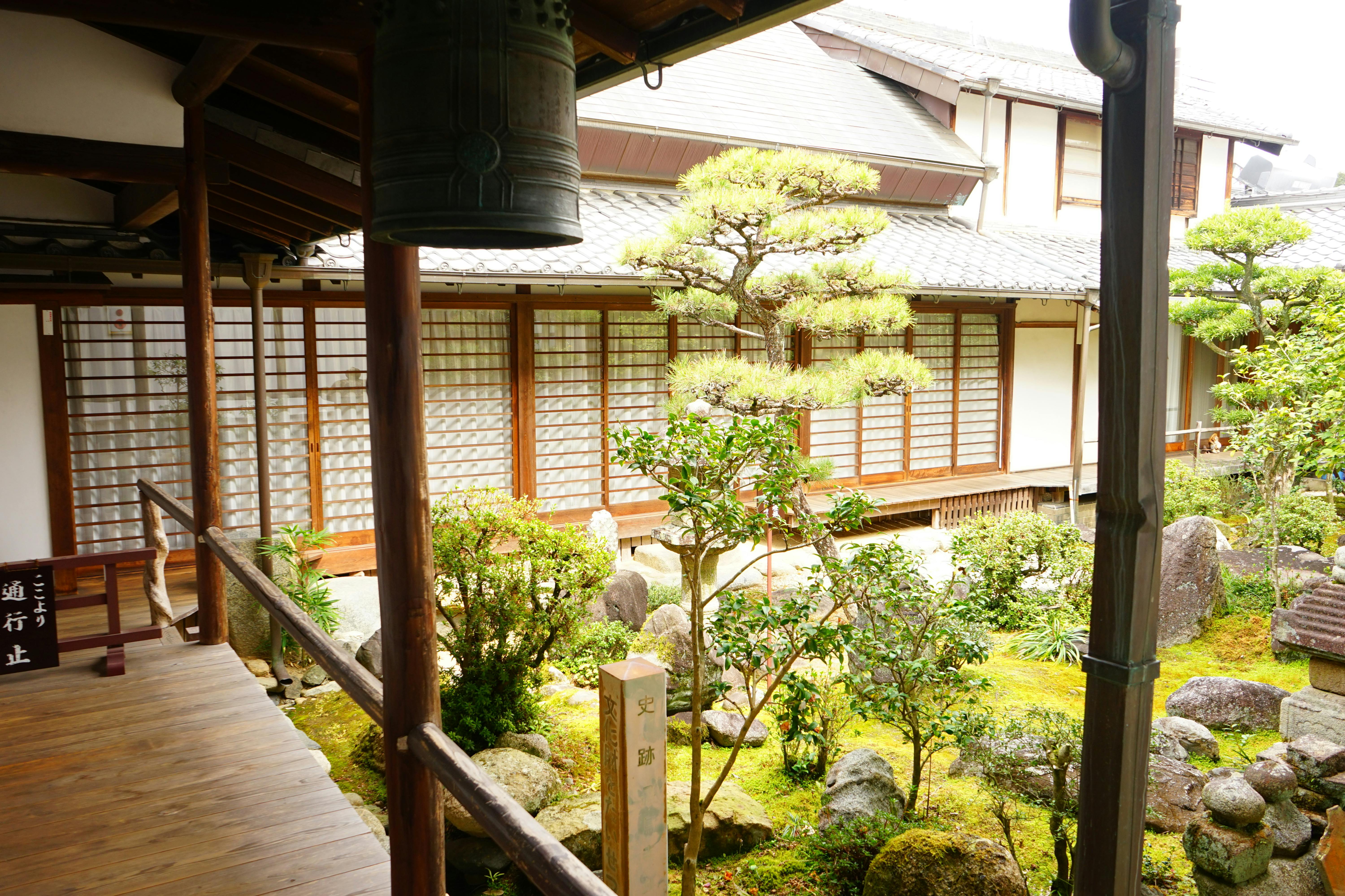 A traditional Japanese courtyard garden with manicured trees, rocks, and moss, surrounded by wooden walkways and shoji screen doors, part of a serene building with a tiled roof.