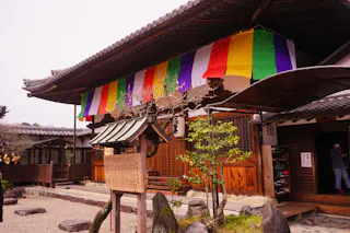 A traditional Japanese building decorated with colorful vertical banners. A small wooden shrine and sign stand in the stone garden in front, with a person partly visible inside the entrance.