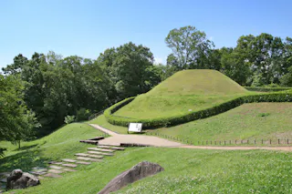 A grassy burial mound surrounded by lush trees and greenery, with a path and steps leading up to it. A signboard stands near the entrance under a bright blue sky.