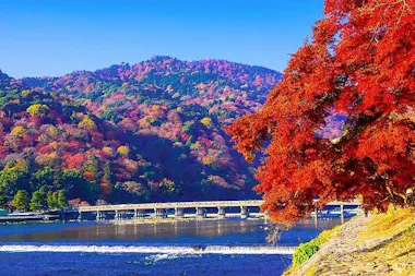 A scenic view of a river with a bridge, vibrant autumn trees with red and orange leaves in the foreground, and multicolored forested hills under a clear blue sky in the background.