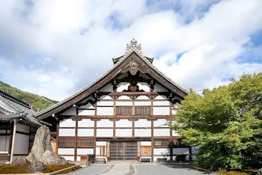 A traditional Japanese temple building with a steep gabled roof, wooden beams, and white walls stands under a partly cloudy sky, with greenery and a large rock in the foreground.