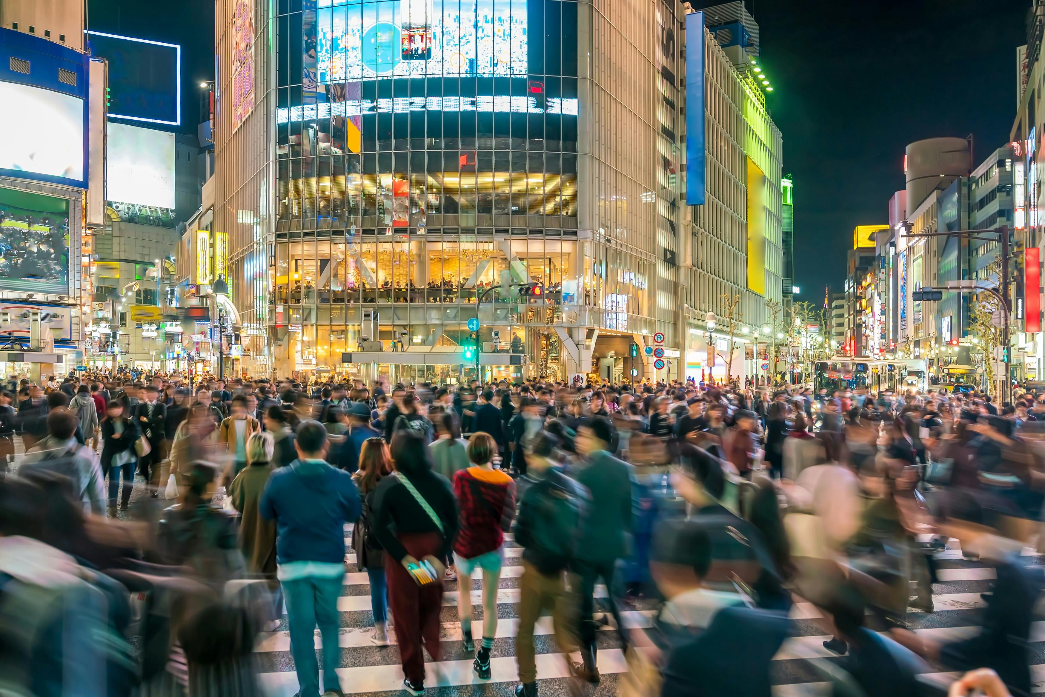 A large, bustling crowd crosses a city intersection at night, surrounded by brightly lit buildings and colorful billboards in an urban setting.
