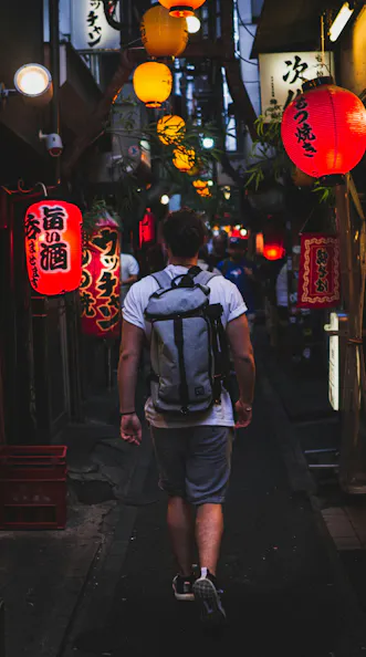 A person with a backpack walks down a narrow alley lined with glowing red and yellow lanterns and Japanese signs, creating a vibrant nighttime atmosphere.