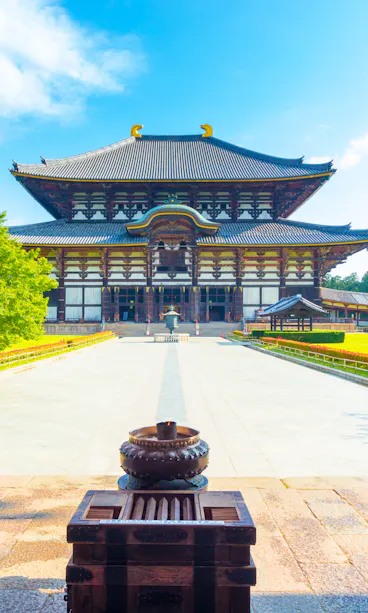Todaiji Temple A traditional Japanese temple with a large wooden roof stands in a sunny courtyard, framed by red pillars and surrounded by green trees and grass under a blue sky.
