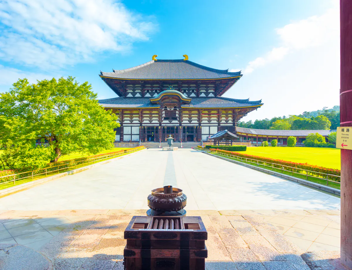 Todaiji Temple A traditional Japanese temple with a large wooden roof stands in a sunny courtyard, framed by red pillars and surrounded by green trees and grass under a blue sky.