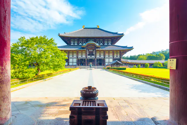 A traditional Japanese temple with a large wooden roof stands in a sunny courtyard, framed by red pillars and surrounded by green trees and grass under a blue sky.