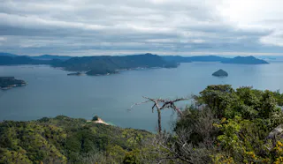 View from a hilltop overlooking a large lake with several islands, surrounded by green hills and dense vegetation under a cloudy sky.